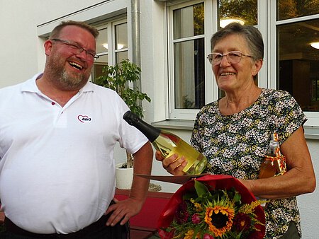 Pflegeheimleiter Thomas Leikam und eine Mitarbeiterin mit Blumen und Sekt lachen miteinander.