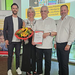 Gruppenbild von vier Personen, drei Männer und eine Frau. Die Frau hält einen Blumenstrauß in den Händen, ein älterer Mann eine gerahmte Urkunde.