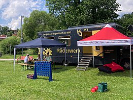 Ein schwarzer LKW mit der Aufschrift "Räderwerk", davor zwei Pavillons mit Spielgeräten.