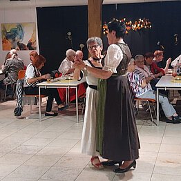 Zwei Frauen tanzen, im Hintergrund sitzen Menschen an Tischen und esssen.