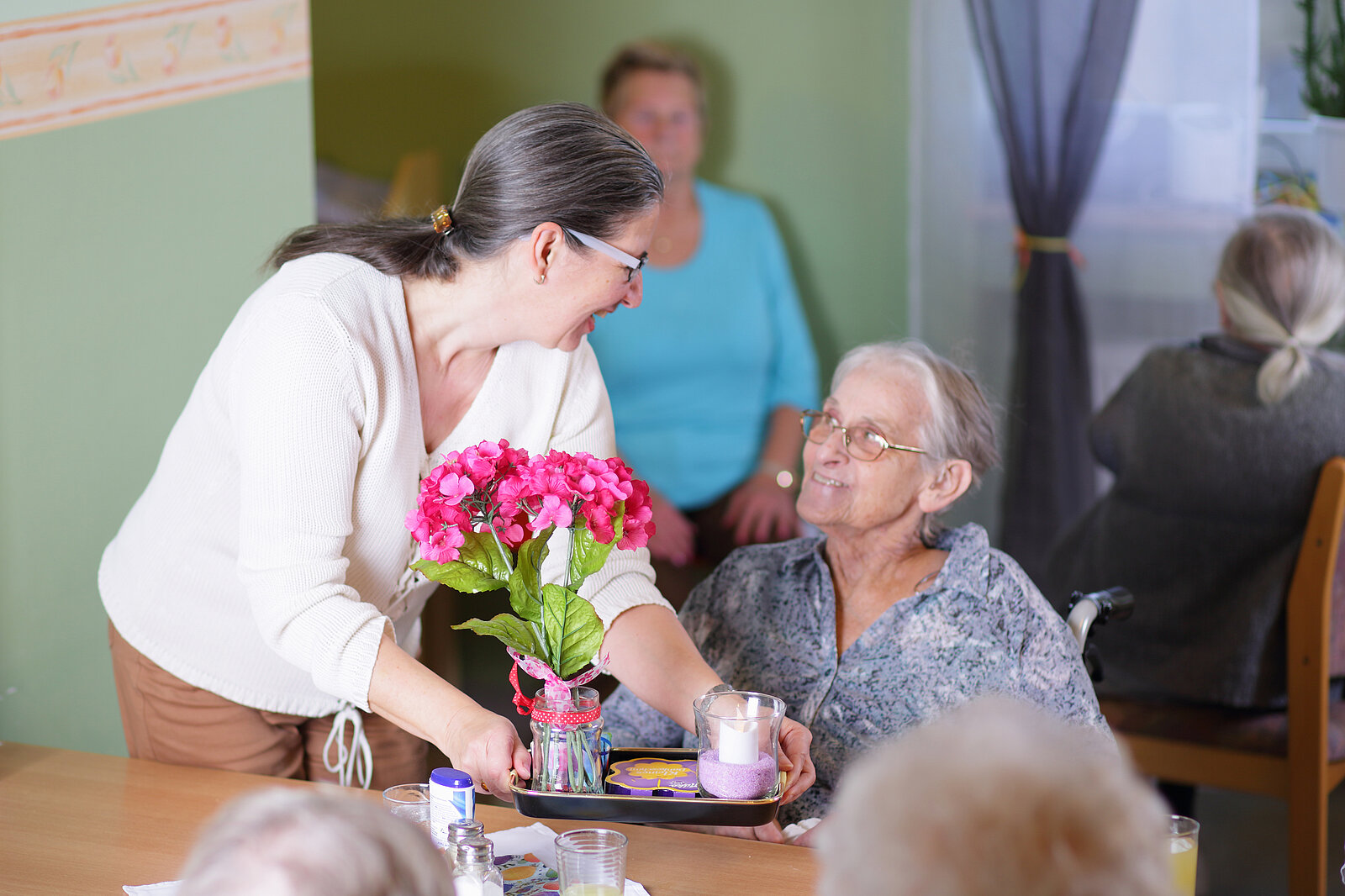 Eine Mitarbeiterin reicht einer Bewohnerin ein Tablett mit einer Tasse und Blumen darauf