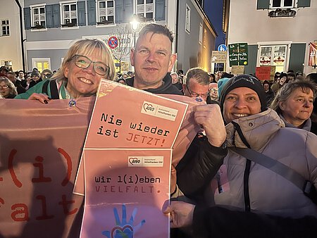 Drei Menschen posieren innerhalb einer Menschenmenge. Sie tragen rosafarbene Plakate, auf denen "Nie wieder ist jetzt" und "Wir l(i)eben Vielfalt" steht.